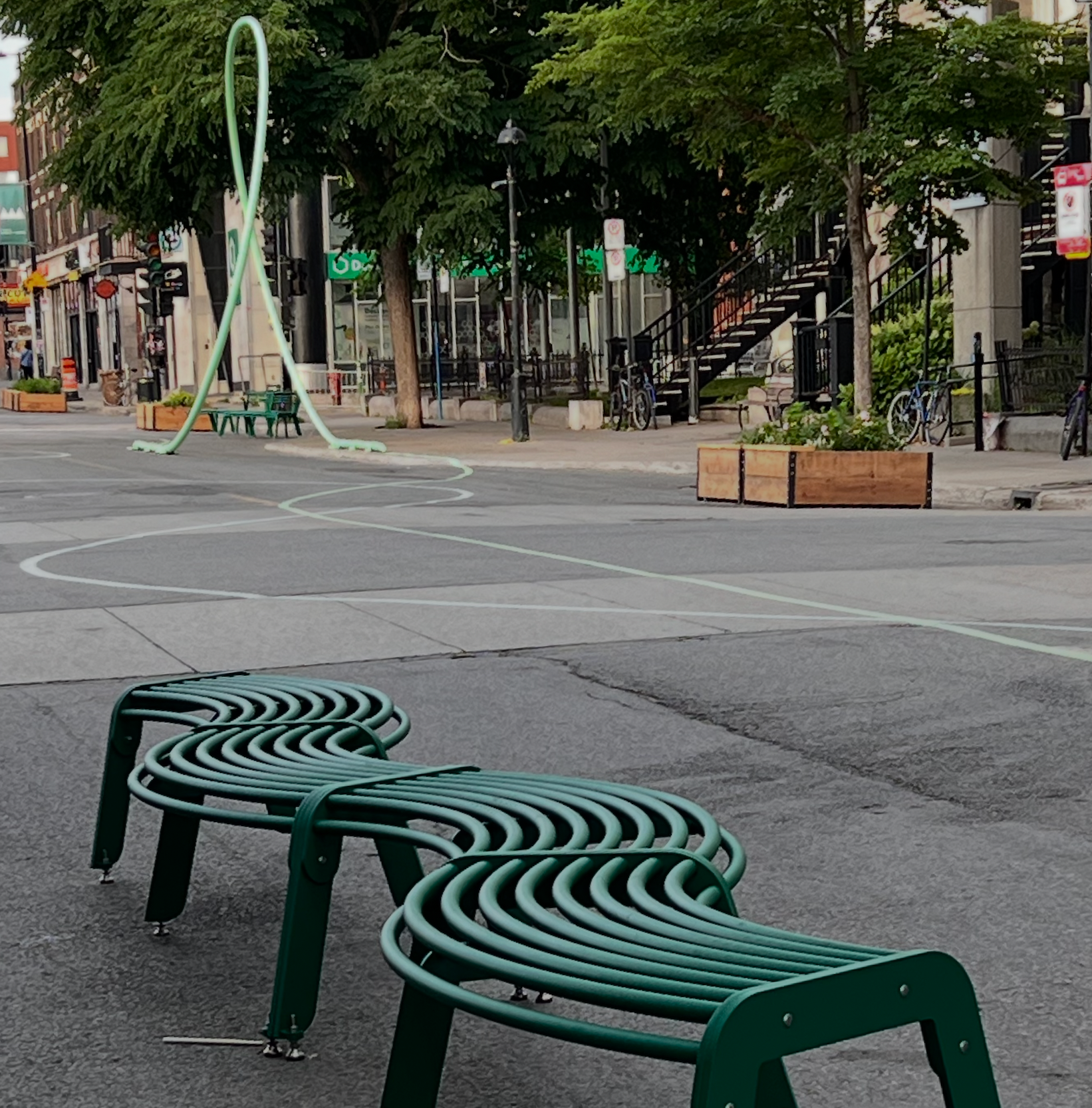 La piéto du Village | banc méandre vert sur la rue avec sculpture tubulaire en arrière-plan
