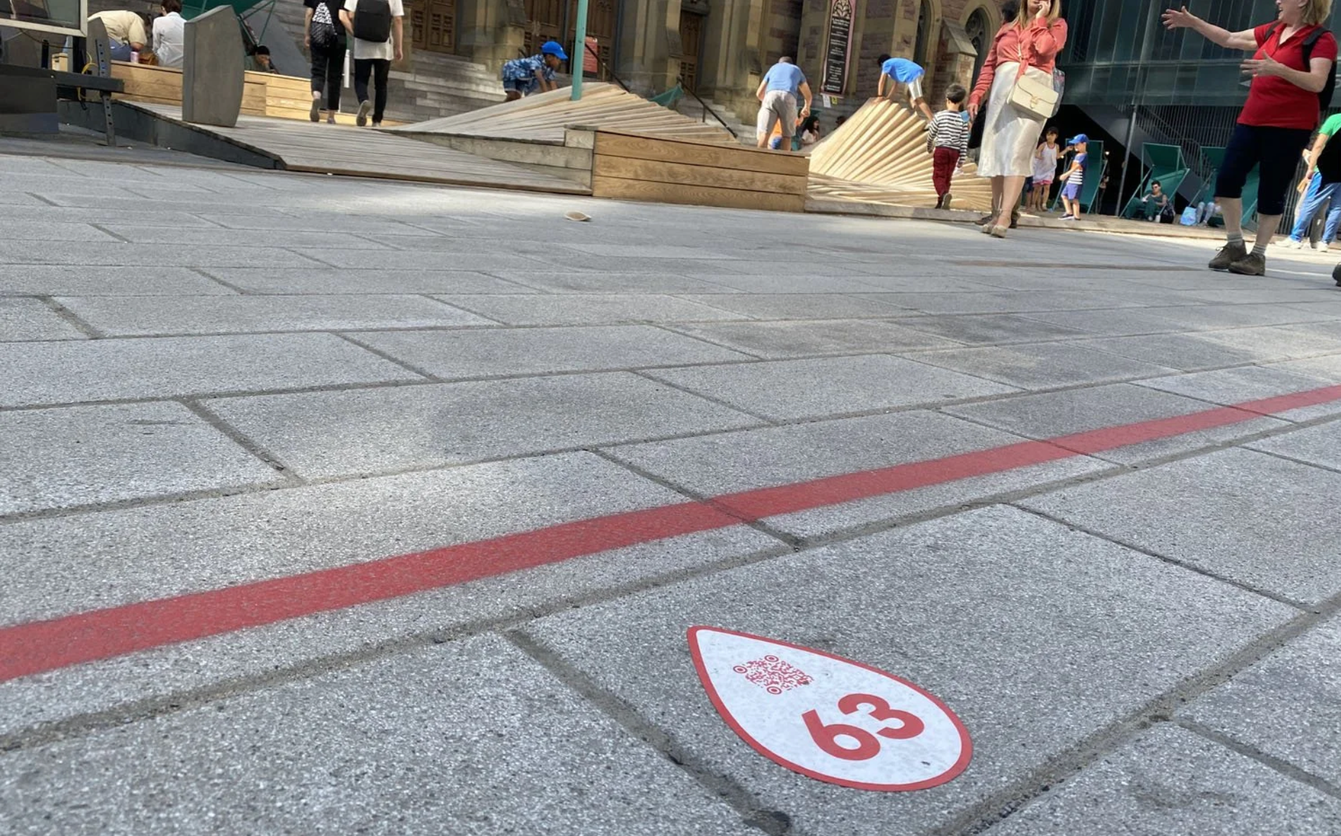 Marqueur n°63 en forme de goutte avec QR code sur le pavé, ligne rouge du sentier et place animée en arrière-plan