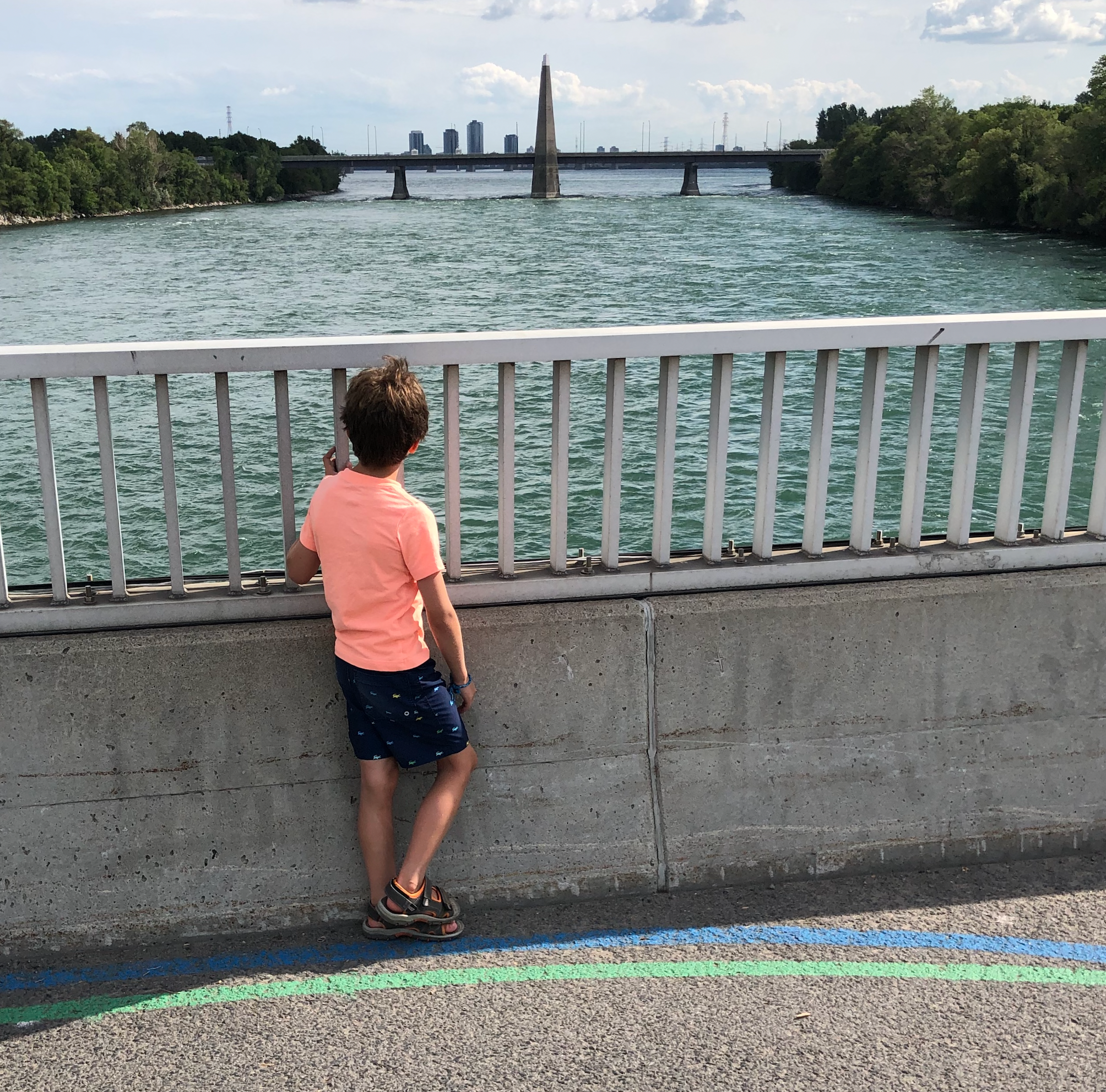 Enfant en t-shirt rose regardant le fleuve depuis un pont avec les tracés vert et bleu au sol