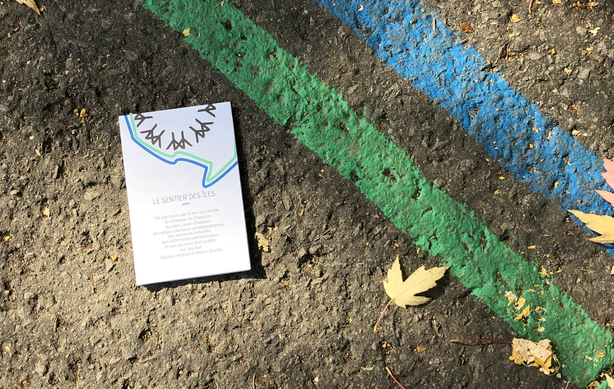 Le Sentier des Îles | brochure posée sur l'asphalte avec les tracés vert et bleu peints au sol